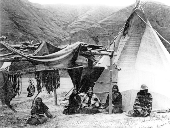 Vintage black and white photo of a family of Shoshone Bannock sitting next to drying racks of fish.