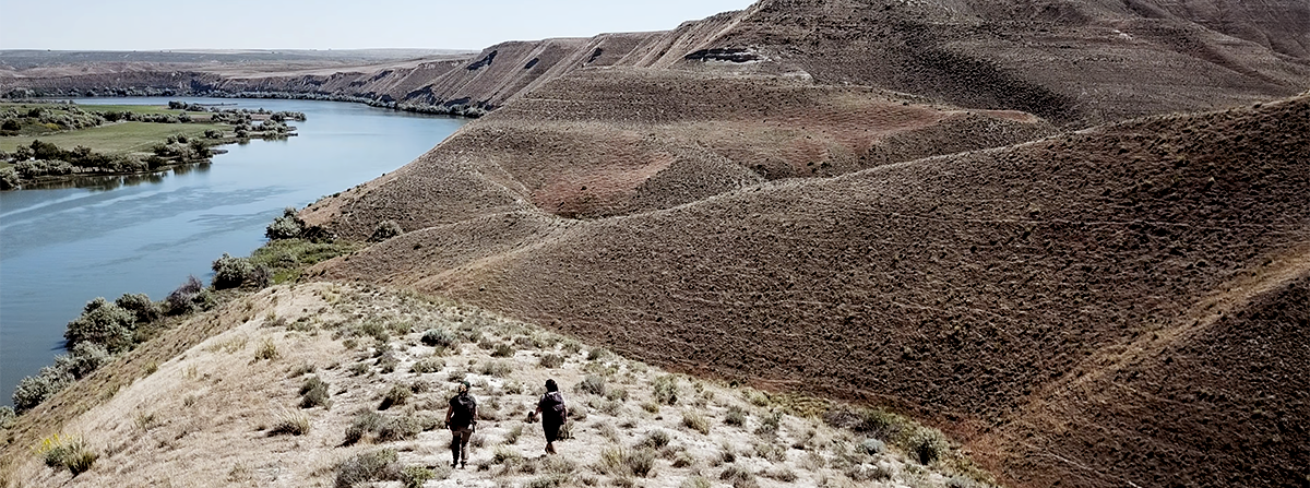 Two small figures walk across the top of the bluffs along the Snake River.