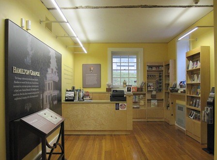 A small room with hardwood floors with a desk, cash register, and shelves with books and other merchandise for purchase, along with an informative exhibit.