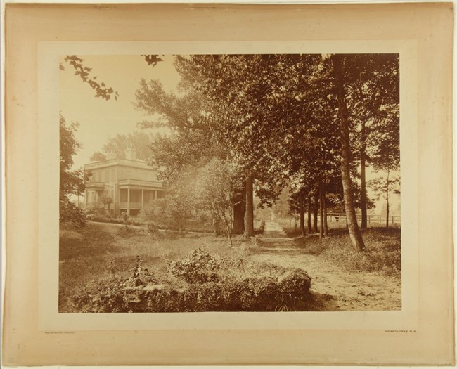 A sepia-tone image of a house surrounded by trees, a dirt road, and grass.