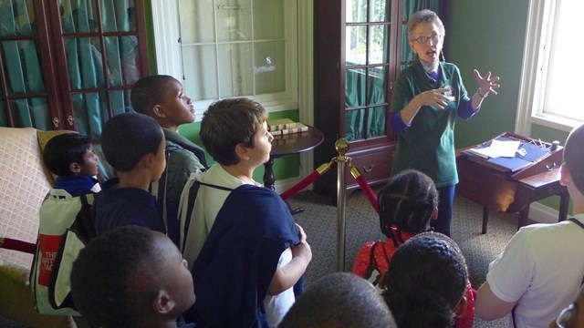 A park volunteer in a green shirt talks to children in a green-walled room.
