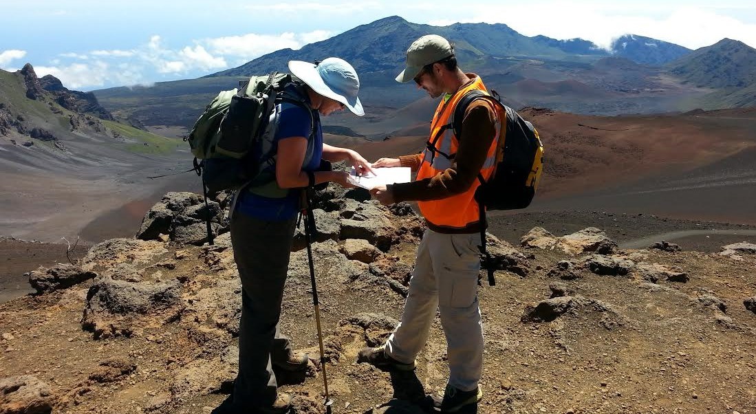 Two people, one in a bright orange vest, stand reading a map along a mountain view trail.