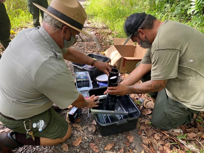 On a rocky trail, two park employees kneel over a configuration of black plastic tubs and tubes to assemble a mosquito trap.