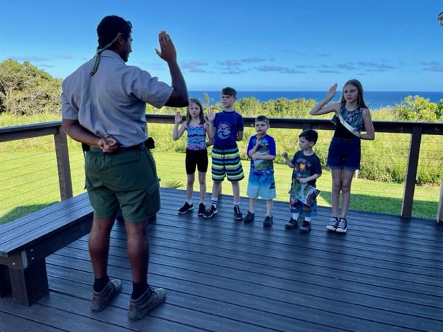 An adult male raising his right hand with three small children facing him on a deck with blue skies, greenery, and ocean in the background.