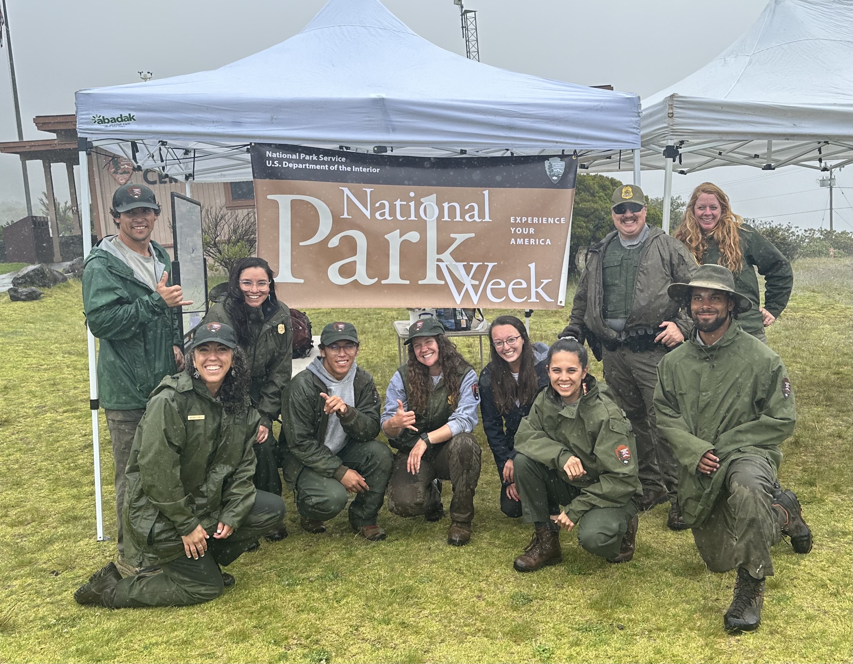 People dressed in green and gray National Park Service uniforms stand outside together in front of a white tent on a rainy day