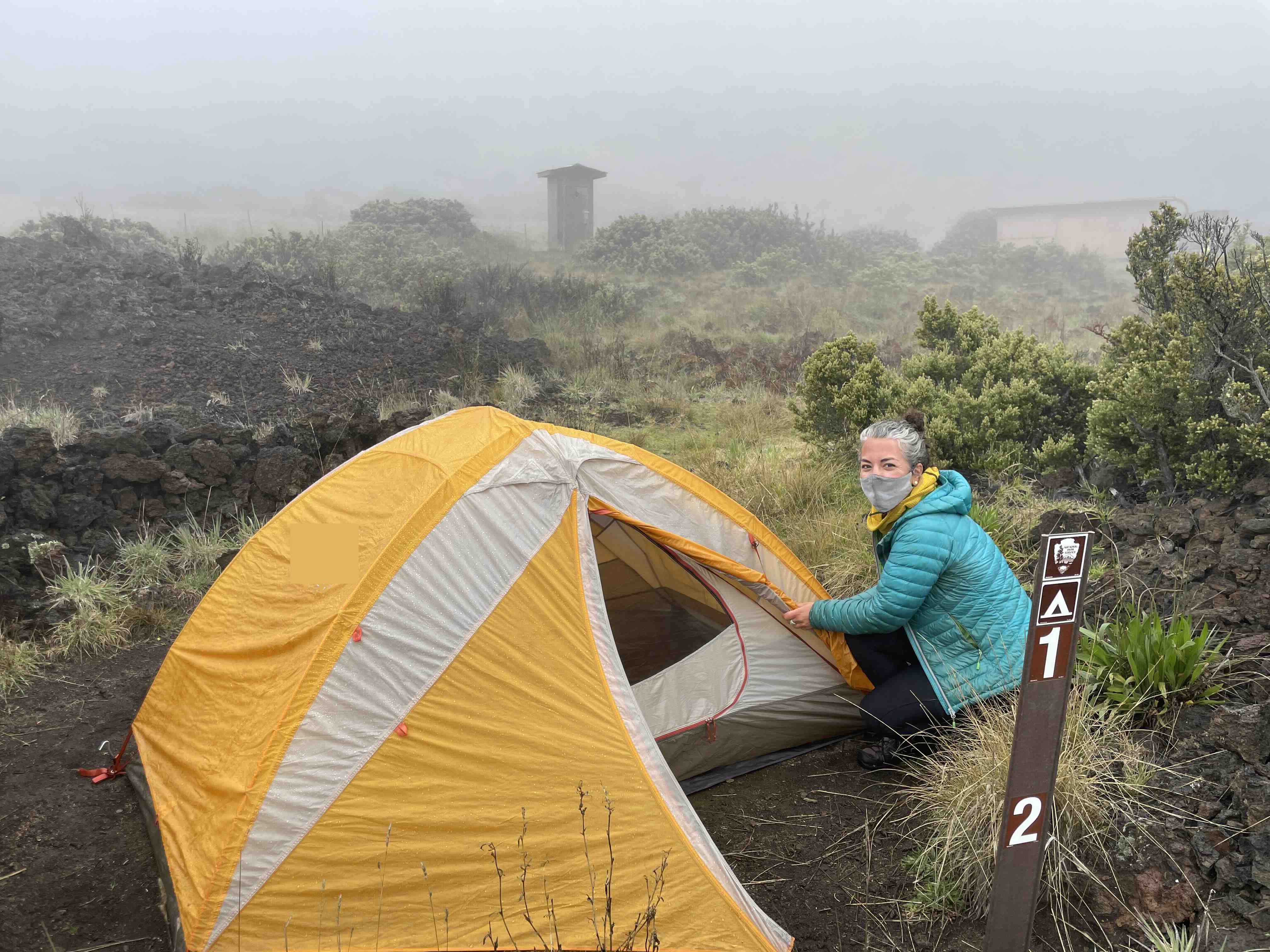Holua Wilderness Campsite with backpacker. person by yellow tent crouched on the ground with marker next to tent