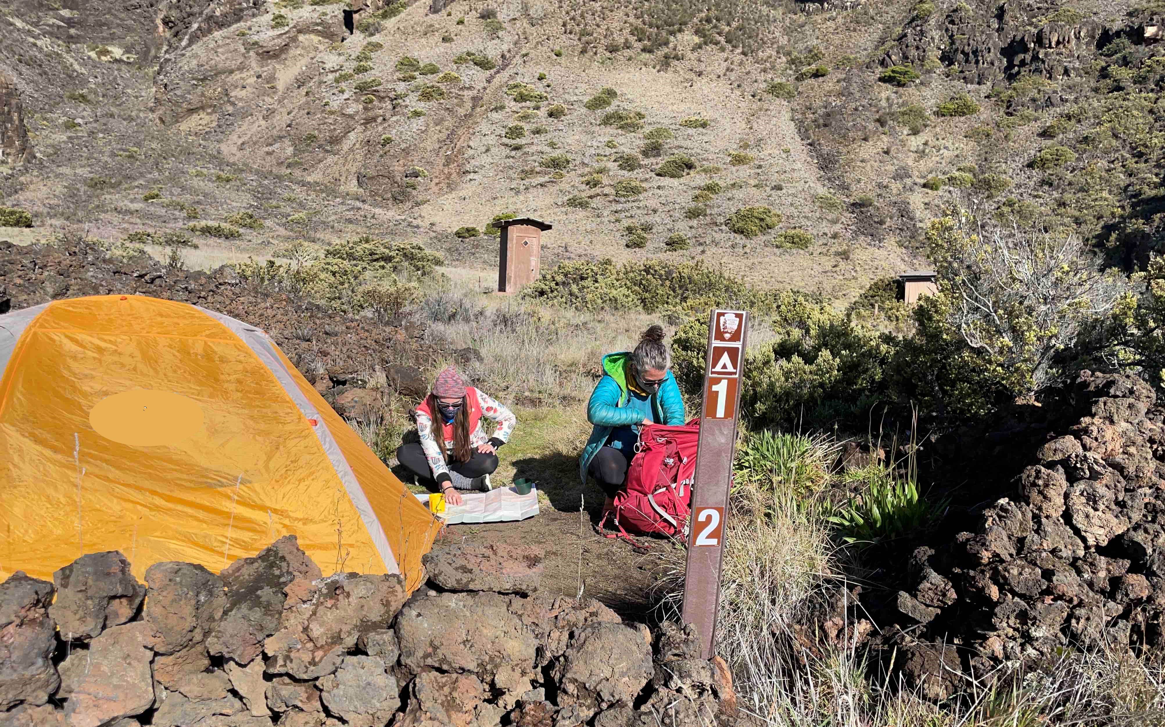 two women next to tent in rocky area with sign post depicting site numbers 1 and 2