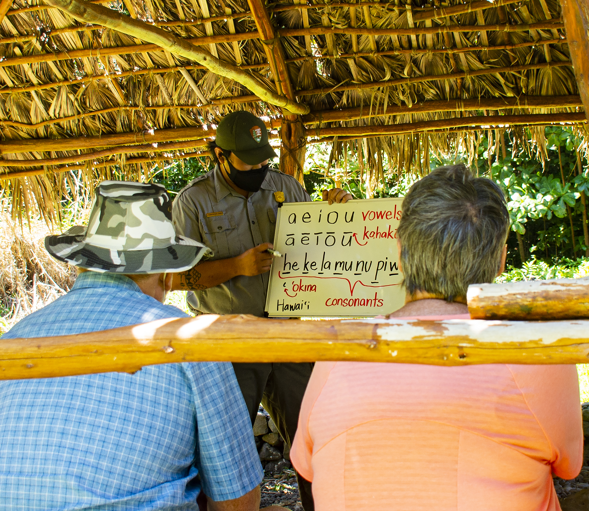 Park Ranger Chris Chow teaches the Hawaiian alphabet to park vistiors in the Halawai in Kipahulu NPS Photo by Jake McFee 4-14-21.jpg person stands with white board in a covered structure teaching letters to two people sitting down