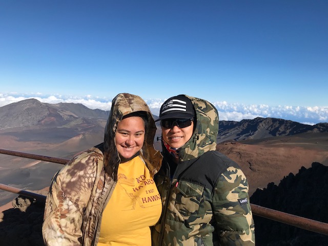 Two women stand at the edge of a railing near a volcanic crater with blue sky above.