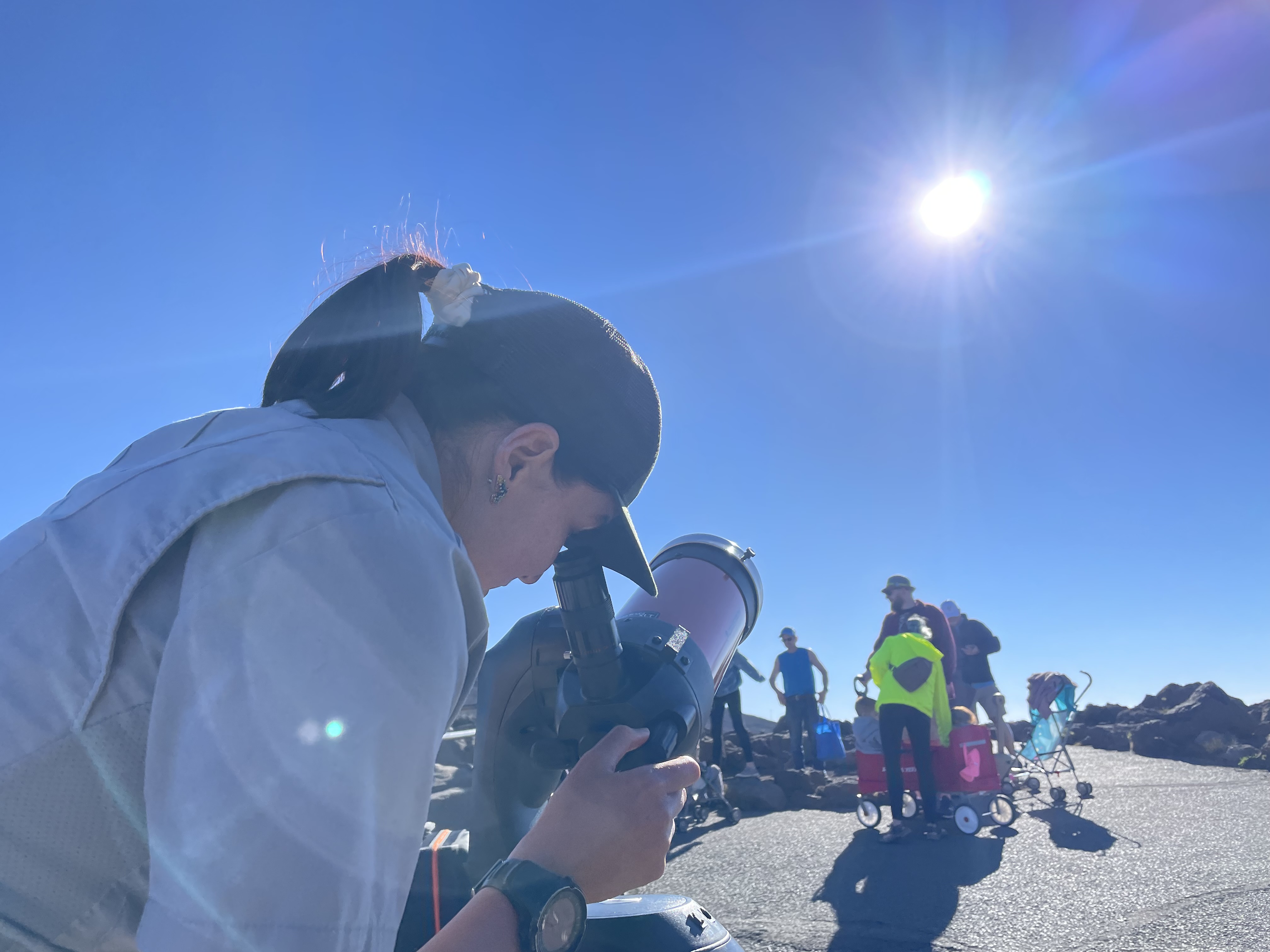 a person with dark hair looks through a white telescope pointed at the sun, blue skies are in the background