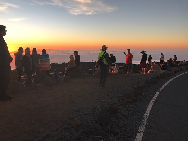 A park employee speaks to a crowd of visitors next to the road as the prepare to watch sunset over the clouds.