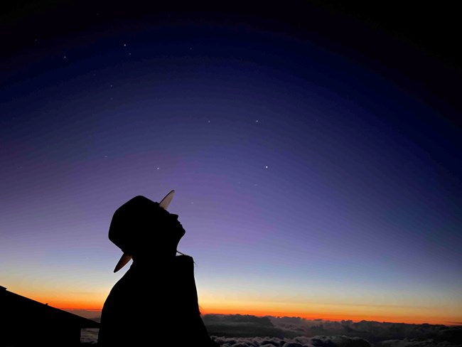 The silhouette of a ranger wearing a flat hat looks up at the stars appearing as the sun sets in the distance.