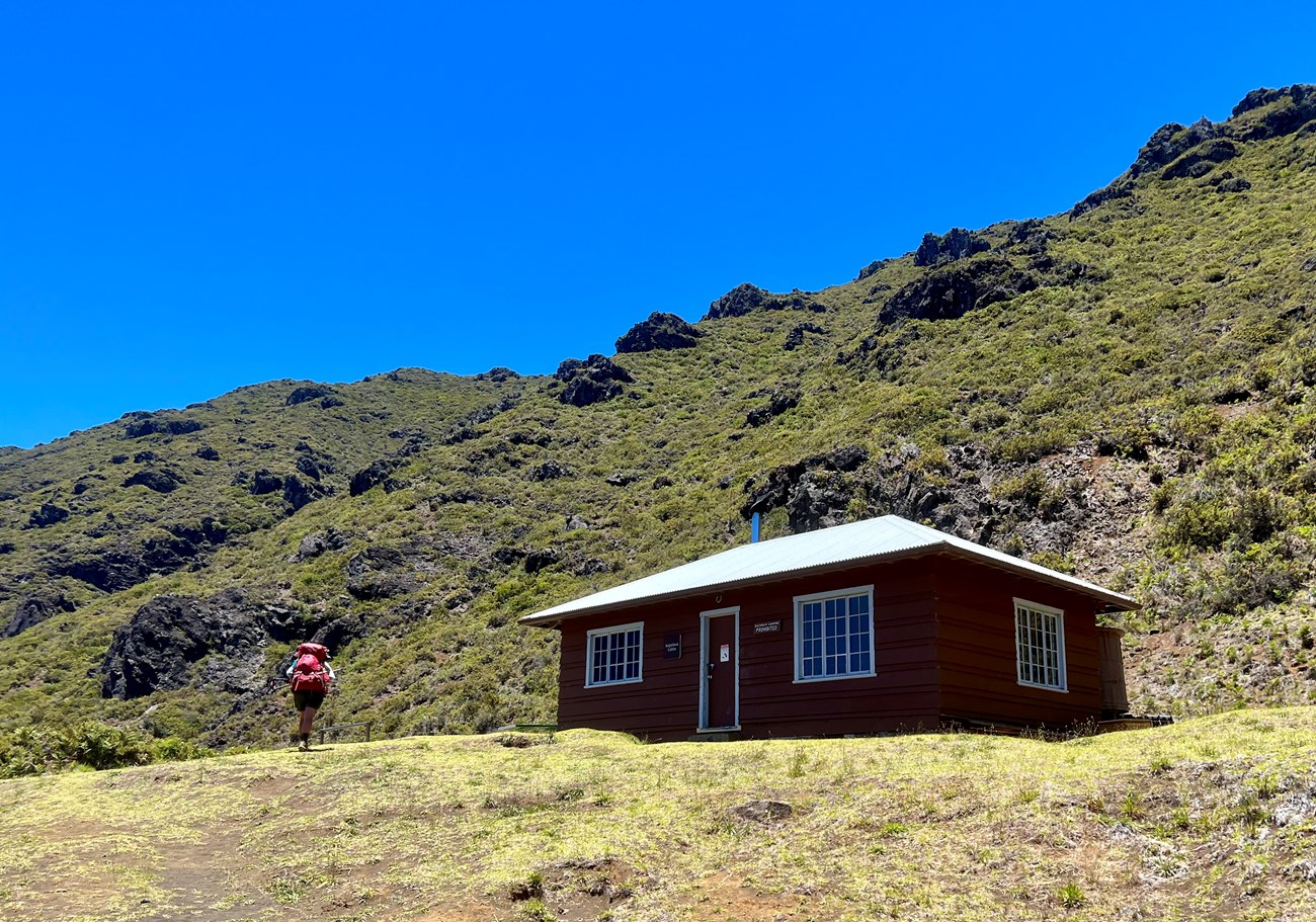 rocky cliff with greenery and cabin in foreground with hiker walking