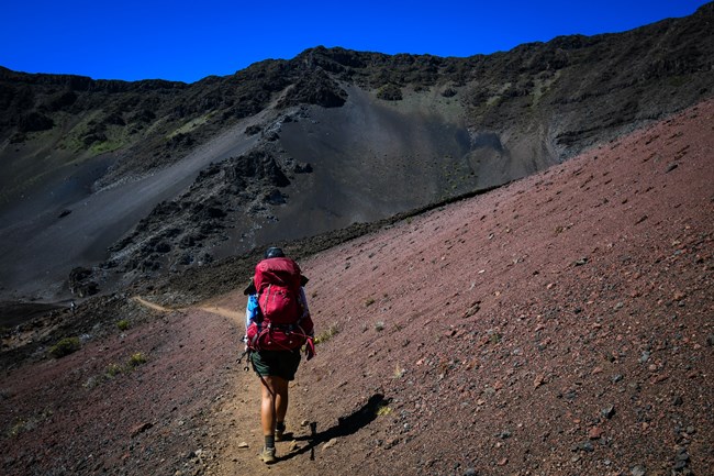 hiker with large red backpack on trail surrounded by red cinder and tall dark peaks in distance
