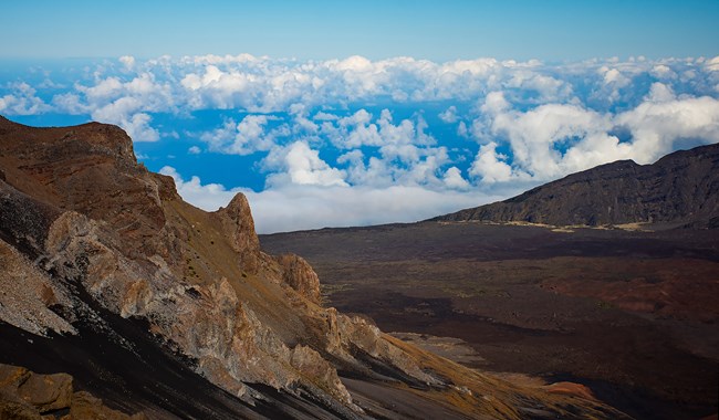 A cloudy and blue sky sits behind a red and brown mountain and crater view.