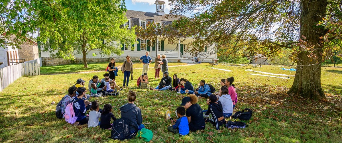 students sit in a circle participating in a field trip program in front of the farm house.