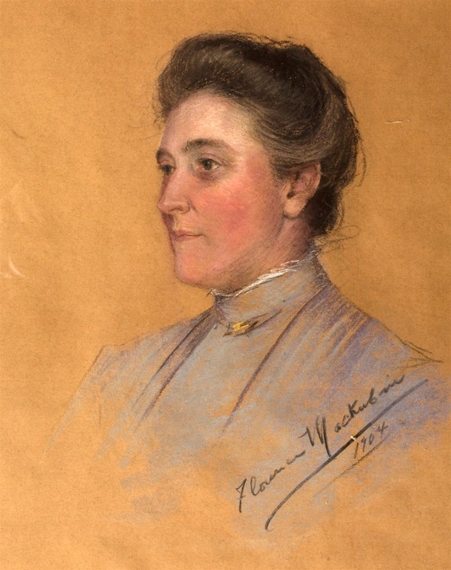 Portrait of a dark-haired lady with hair in a bun and pink cheeks.  She is wearing a gray simple dress.
