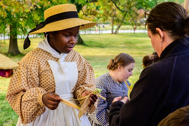 a living historian is holding a cornhusk that they've folded to look like a doll and showing visitors how to do it.