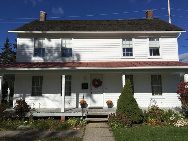 White, two-story house with thin columns on the front porch with shrubbery, orange mums on the porch by the door, and a wreath on the door.