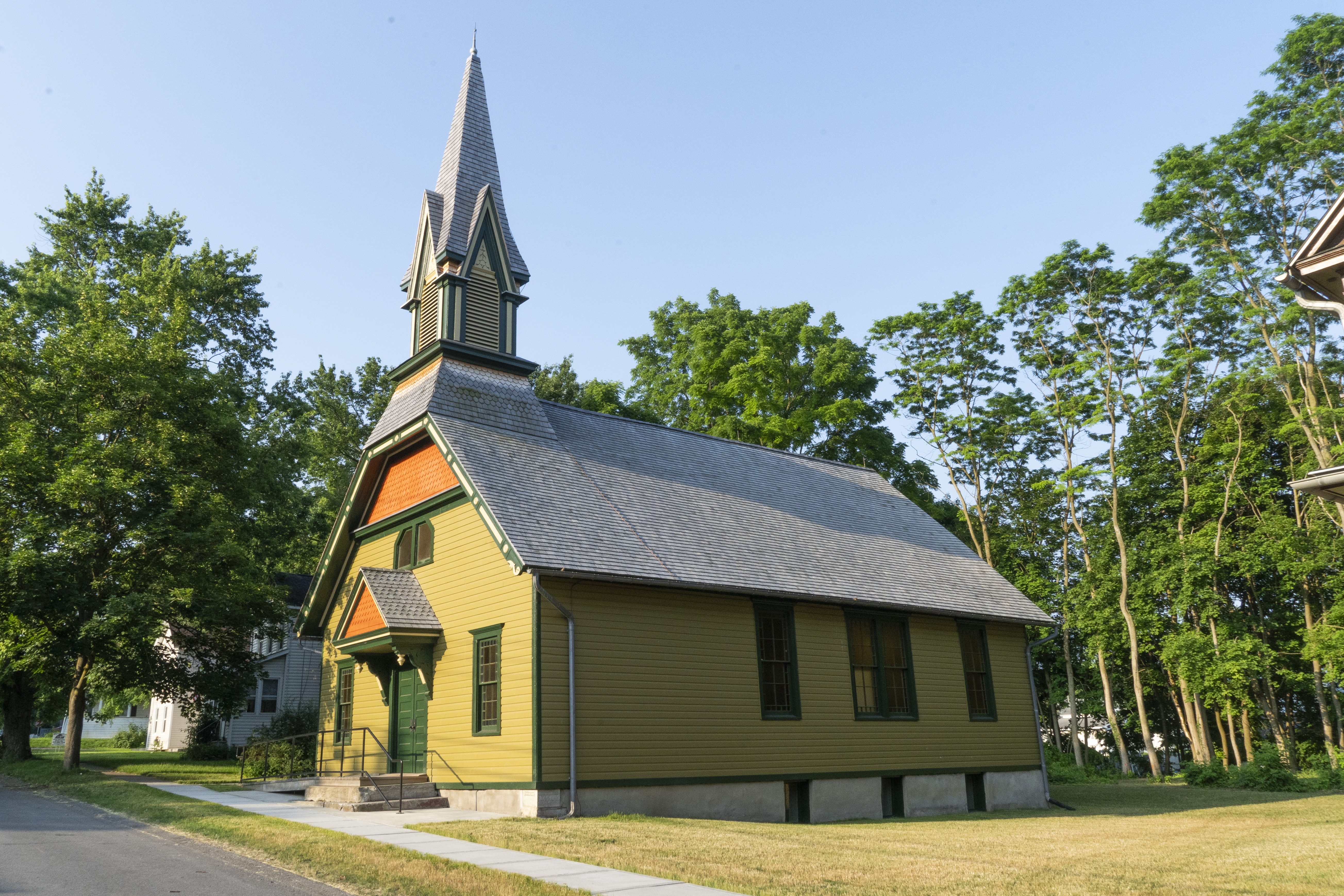 An olive-green colored church in a grassy lawn, framed by trees.