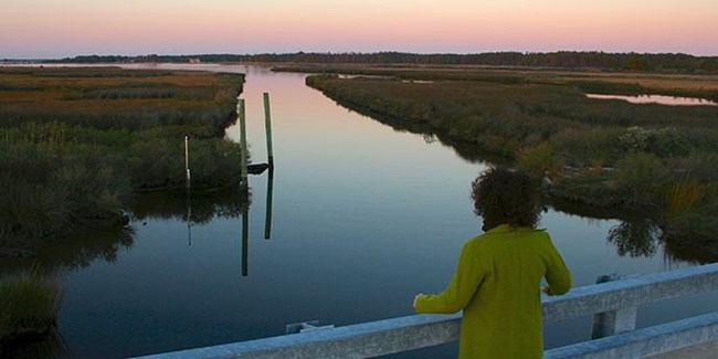 Sunset along Stewart's Canal, with a woman standing on a bridge overlooking the straight corridor of water with pink and purple skies