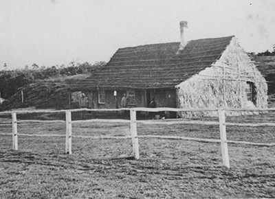 Black and white photo of a grass-thatched building behind a wooden fence with two small human figures standing on the porch