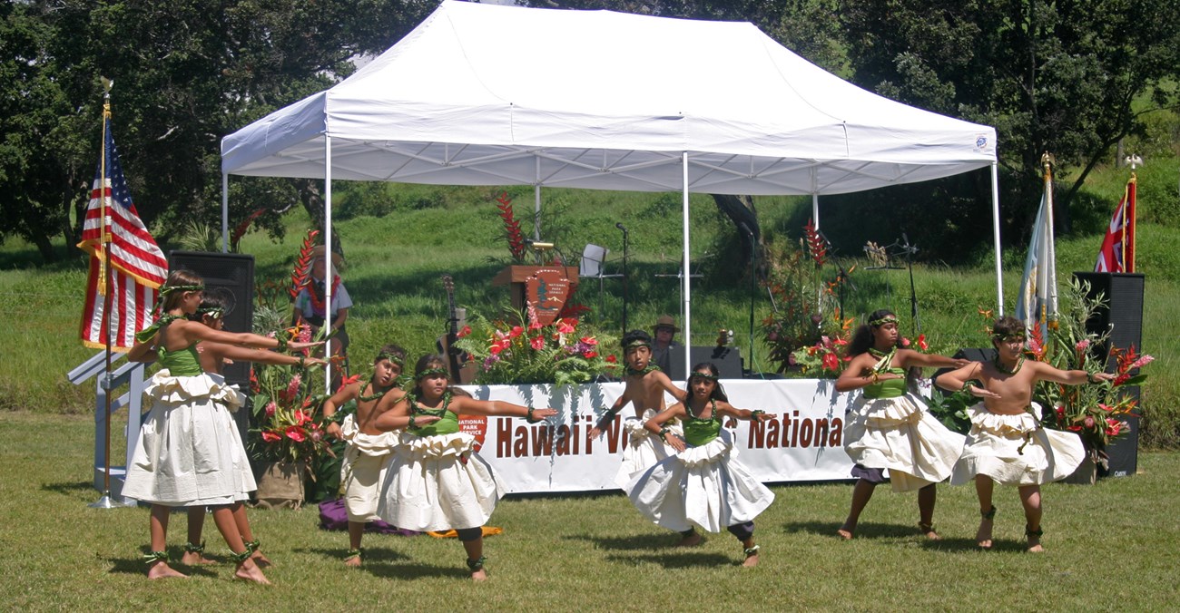 A halau hula of children in front of a white tent and stage