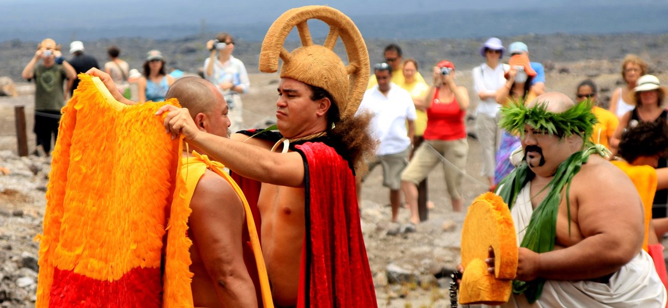 Aliʻi reenactor placing a cloak on another while a third assists