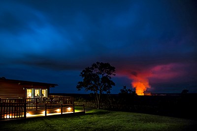 Building lit with electric light at night, on the edge of a volcanic crater with an orange glowing plume in the distance