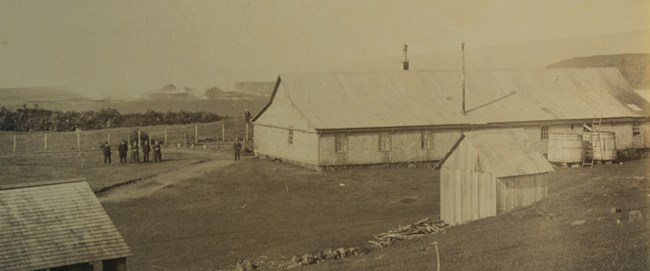 Black and white photo of a large wooden building and two sheds overlooking a volcanic crater