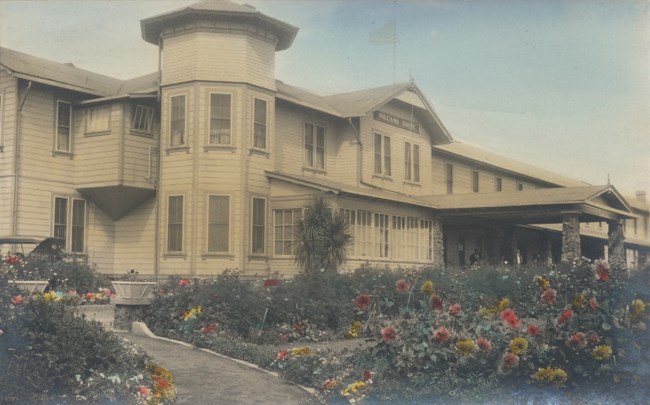 Hand-colored photograph of the Volcano House hotel, a large white building with flower gardens in front