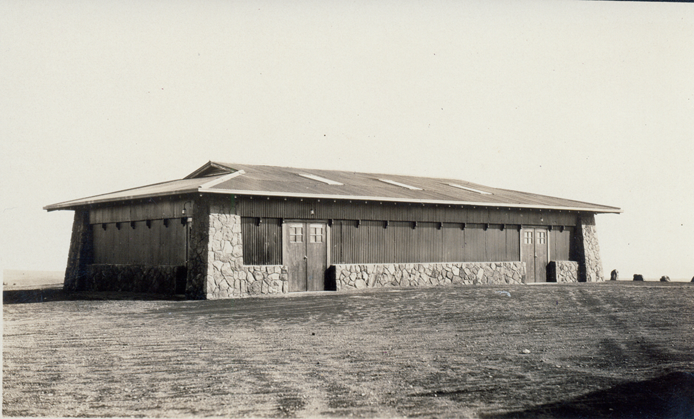 Black and white image of one-story rectangular building with lava rock foundation.