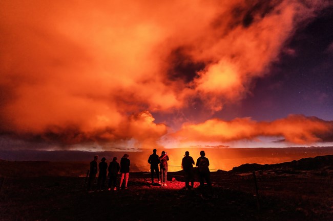 People standing at a cliff overlook at night with bright orange light glowing from a crater and orange clouds of smoke.