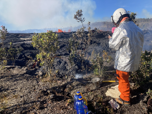 Geologists put molten lava into a metal bucket and rapidly quench it with water. Figure in silver and orange protective gear gathering a lava sample next to dark new lava rock and bright red fountaining lava in the distance.