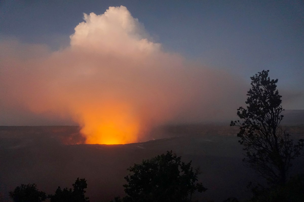 Glowing plume from an erupting volcanic caldera with tree silhouettes in the foreground