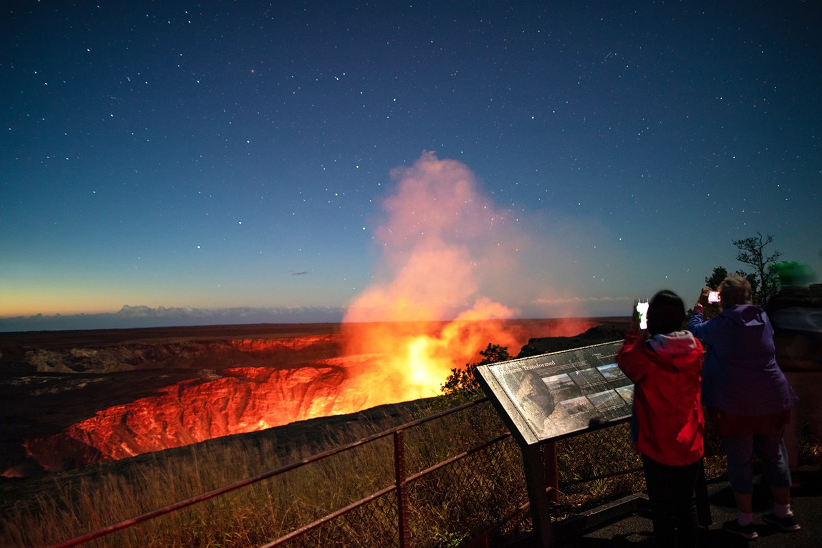 Two people standing at an overlook toward an erupting volcanic crater at night