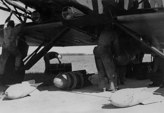 Black and white photo of three bombs on the ground next to a plane and the back of two people in flight suits
