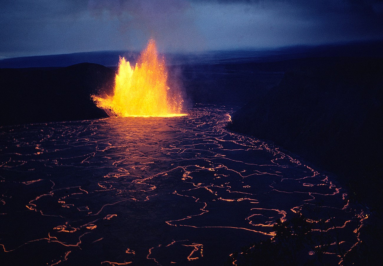 Lava lake in a volcanic crater at dusk in front of a glowing lava fountain and cinder cone