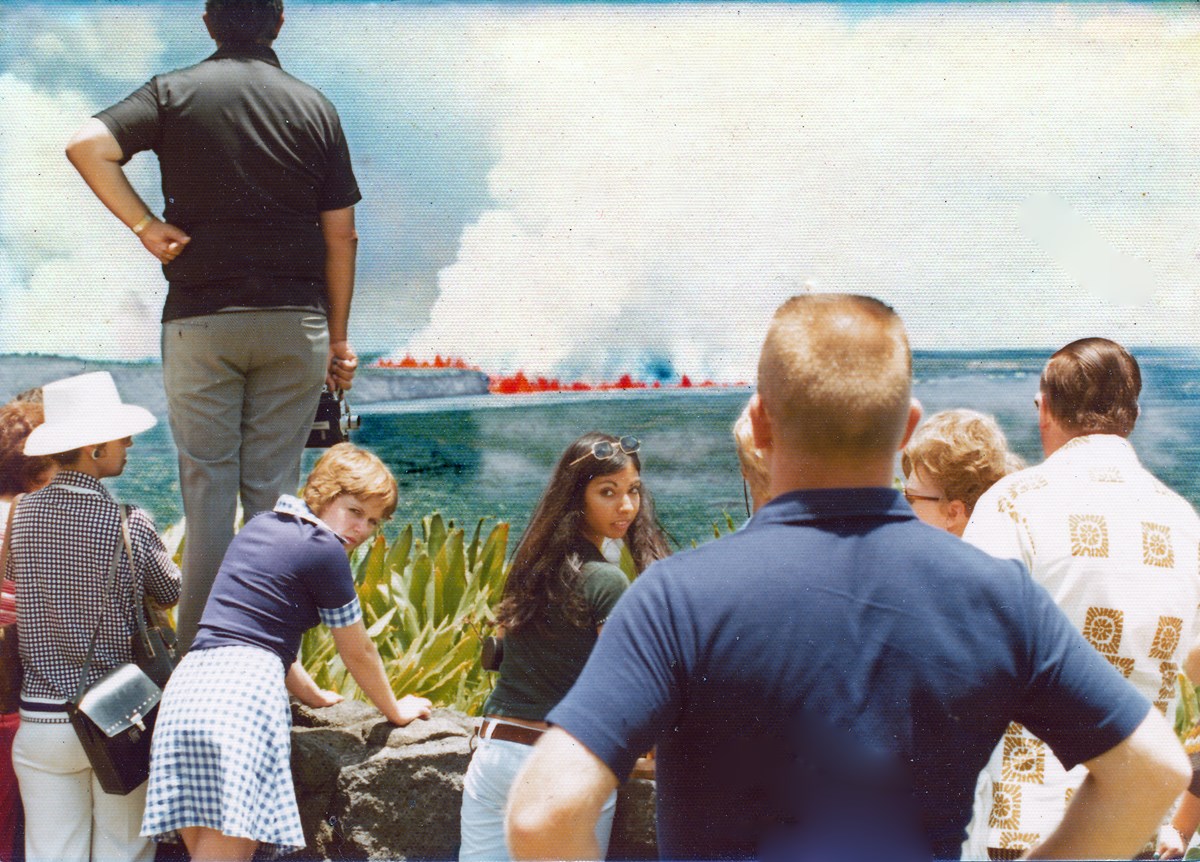 A group of people, including a woman looking backward, at an overlook facing a volcanic caldera and distant lava fountains