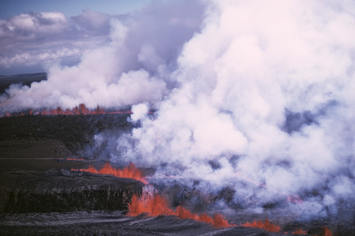 Aerial view of lines of erupting lava fountains sending out fumes and cascading lava into a pit crater and larger caldera