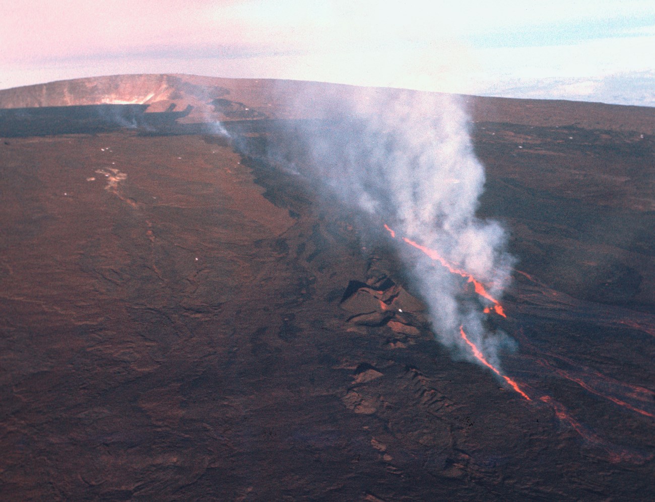 Aerial view of a volcanic fissure spewing lava with a large volcanic caldera in the background