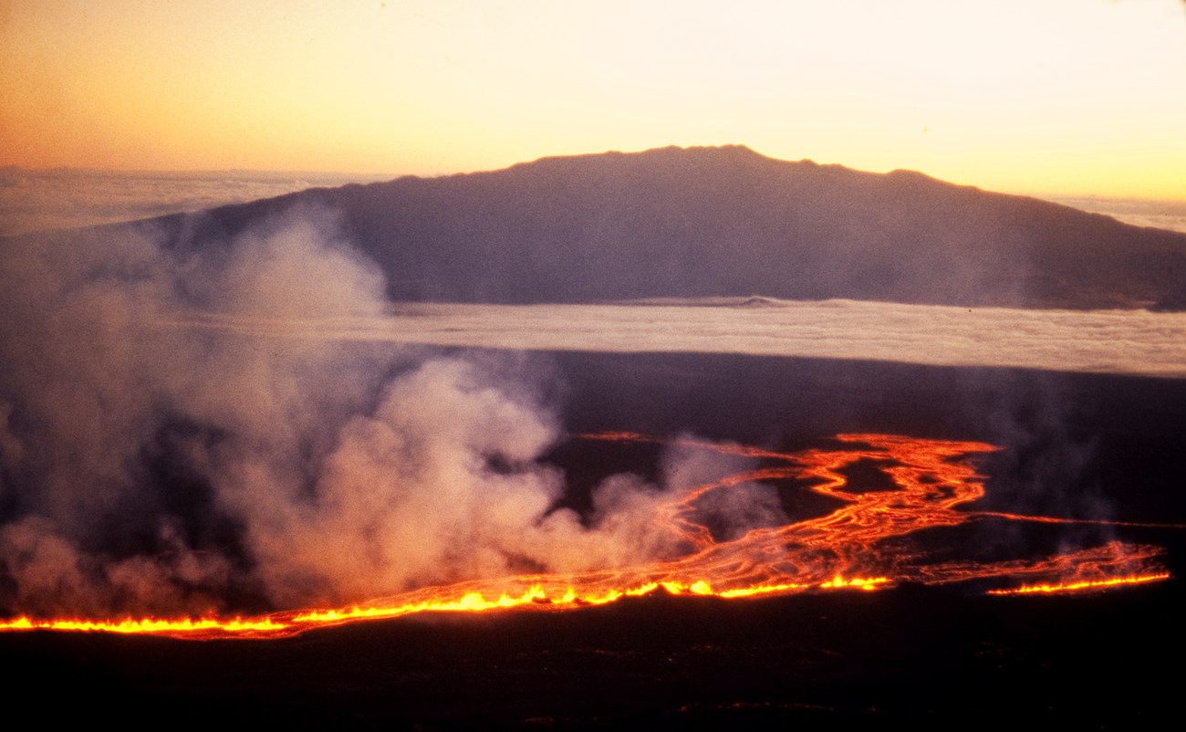 Molten lava flows on a mountainside with a silhouetted mountain beyond at sunrise