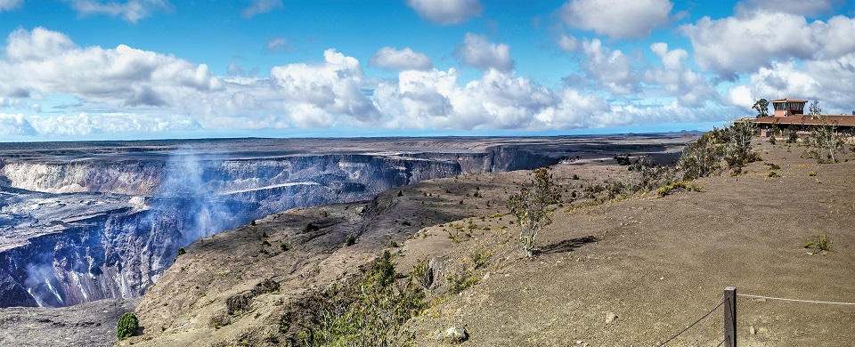 deep cracks and crumbling cliffs at the top of a volcano crater, with a building sitting close the edge of the crater