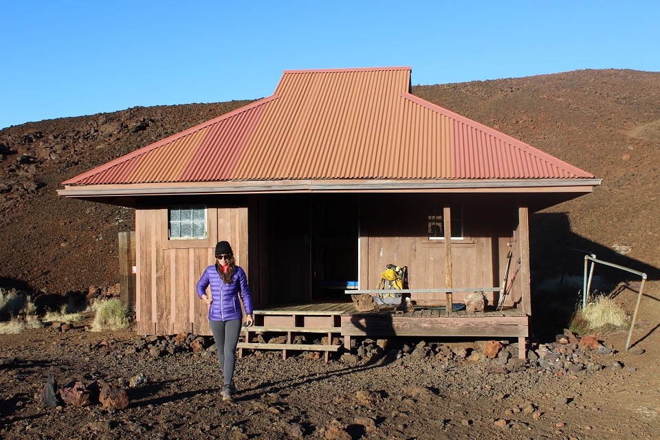 A hiker in cold weather garb on the deck of a rustic red cabin