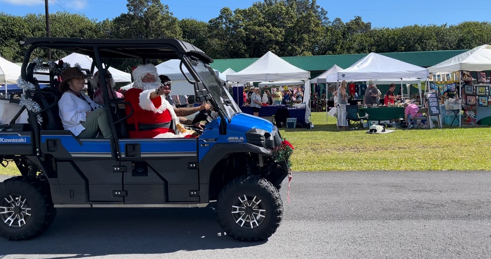 960-FHVNP - Holidays at Kahuku Santa & crafters 2023 Santa Claus rides shotgun in a blue all terrain vehicle with passengers