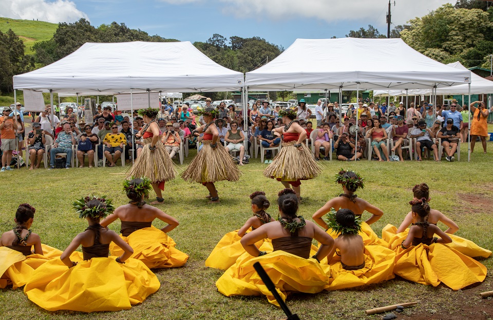 Young hula performers dance for a crowd of people sitting under a white tent