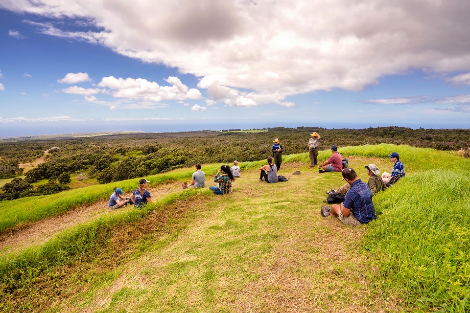 A group of people sitting atop at grassy hill in the sunshine, with the ocean in the distance.