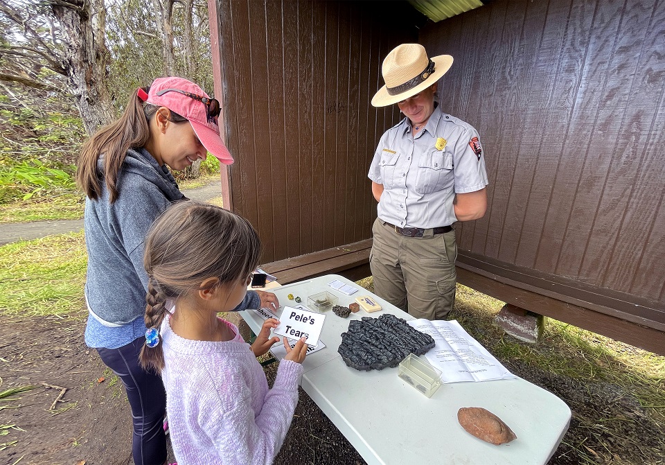 960-HAVO_202304_Ranger Jody Anastasio and keiki at NP Week event_J.Wei A park ranger engages with a young girl who holds a sign that says Peleʻs Tears