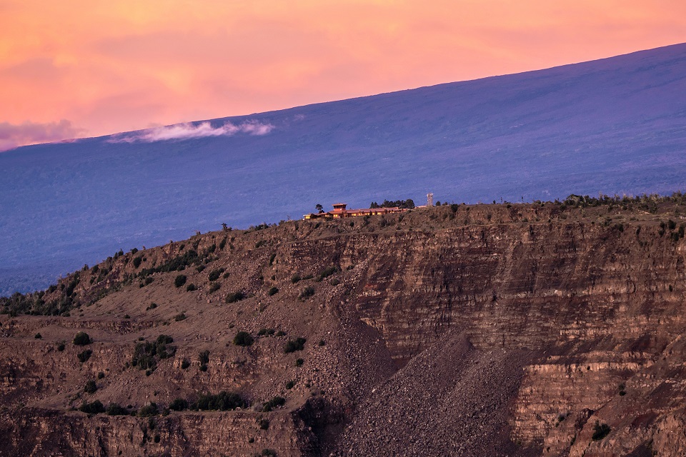 Buildings on the edge of a steep volcanic crater with a mountain looming above them