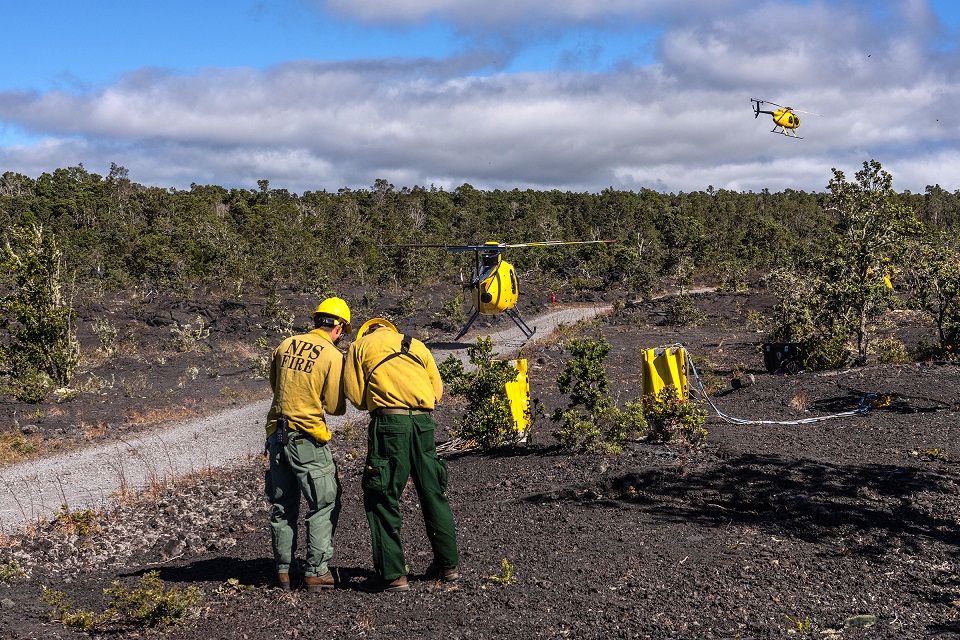 960-HAVO_20241009_NPS-firefightera-at-helicopter-buckets-Makaopuhi-Fire_J.Wei Two NPS firefighters confer as two yellow helicopters take off